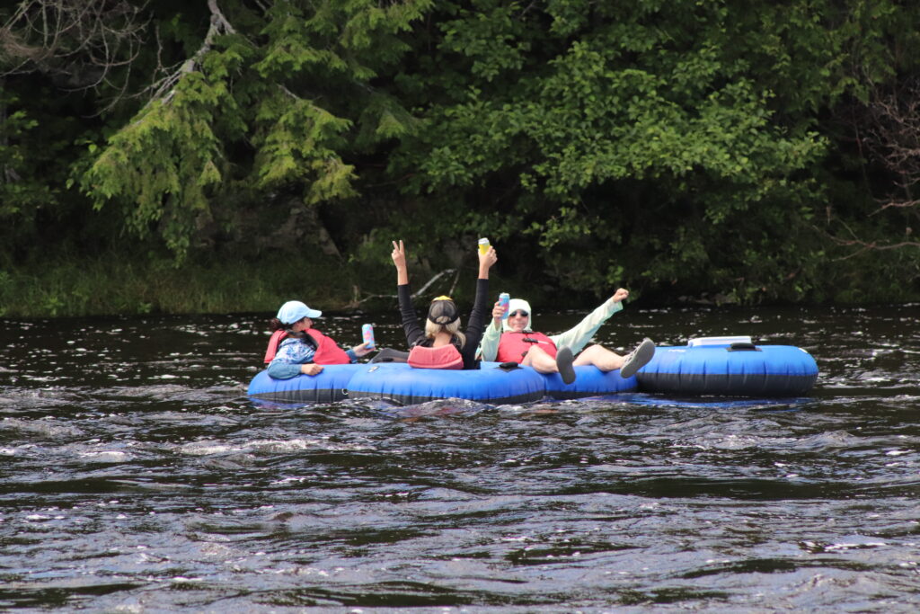 Three people sitting in blue tubes on the Kennebec River, cheering as they float by.